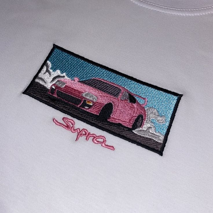 Rosafarbener Toyota Supra mit Schriftzug Supra, gestickt auf weißem Shirt